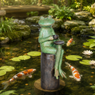Frog statue with a cup on a stone in a pond with koi fish and lily pads.